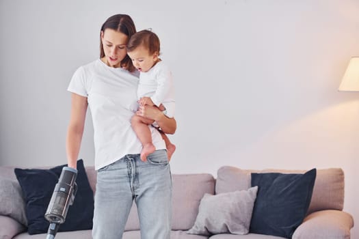 Woman uses vacuum cleaner. Mother with her little daughter is indoors at home together.