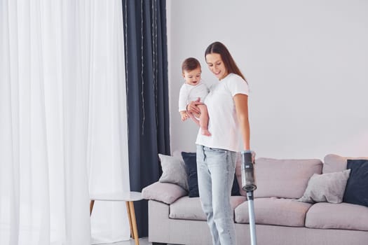 Woman uses vacuum cleaner. Mother with her little daughter is indoors at home together.