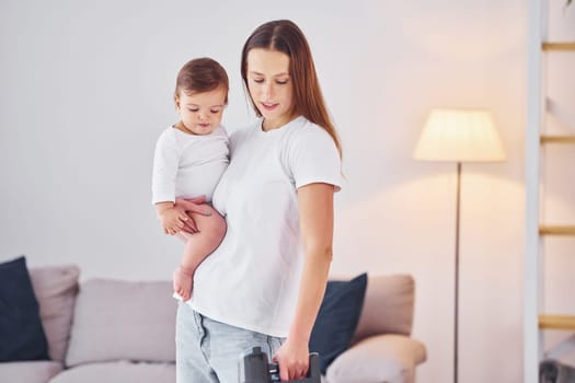 Woman uses vacuum cleaner. Mother with her little daughter is indoors at home together.