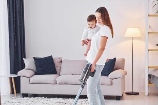 Woman uses vacuum cleaner. Mother with her little daughter is indoors at home together.