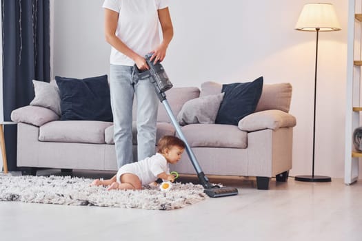 Using of the vacuum cleaner. Mother with her little daughter is indoors at home together.