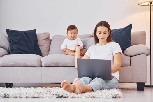 Woman working by using laptop. Mother with her little daughter is indoors at home together.