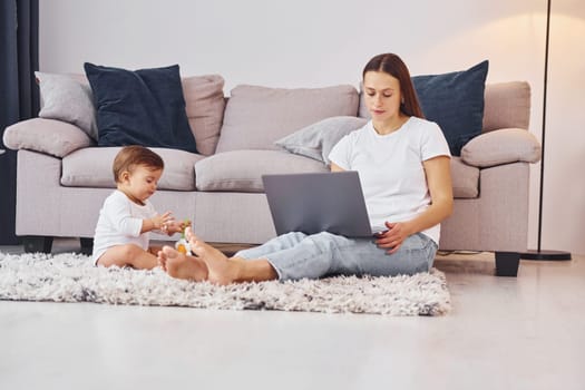 Woman working by using laptop. Mother with her little daughter is indoors at home together.