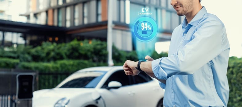 Progressive businessman check wearable hologram watch on electric car battery that being recharge at charging station point in the city. Digital interface, data from EV car display on smartwatch.