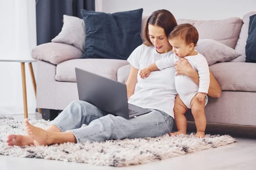 Woman working by using laptop. Mother with her little daughter is indoors at home together.