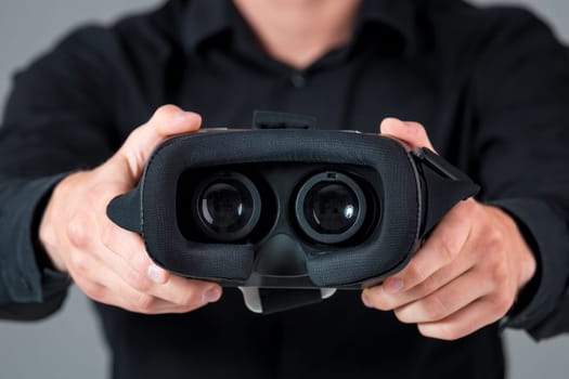 Excited young man using a VR headset and experiencing virtual reality on grey blue background. He holds glasses for viewer