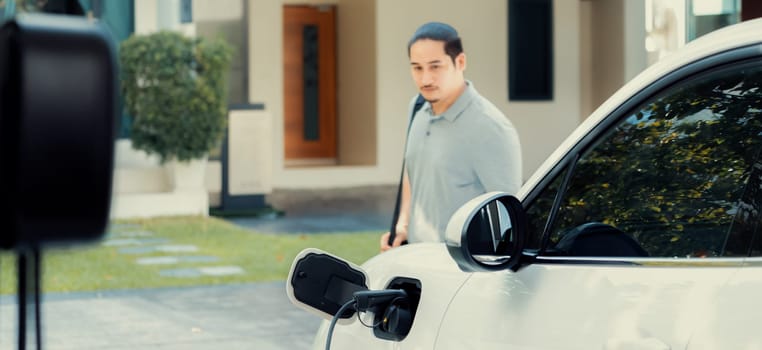 Progressive asian man install cable plug to his electric car with home charging station in the backyard. Concept use of electric vehicles in a progressive lifestyle contributes to clean environment.