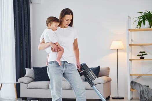 Cleaning floor by using vacuum cleaner. Mother with her little daughter is indoors at home together.