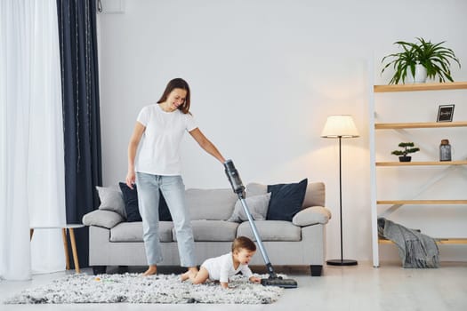 Cleaning floor by using vacuum cleaner. Mother with her little daughter is indoors at home together.