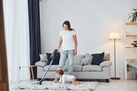 Cleaning floor by using vacuum cleaner. Mother with her little daughter is indoors at home together.