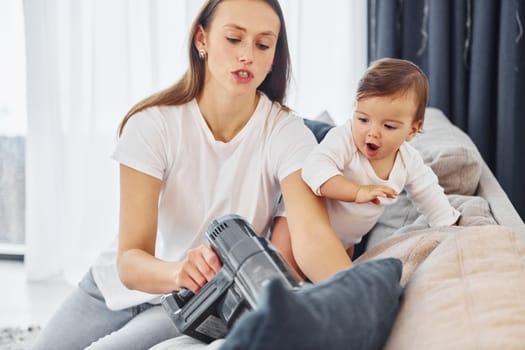 Holding vacuum cleaner. Mother with her little daughter is indoors at home together.
