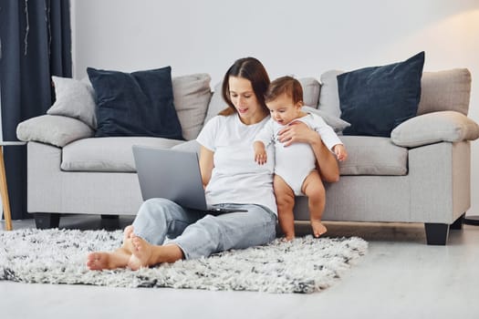 Silver colored laptop. Mother with her little daughter is indoors at home together.