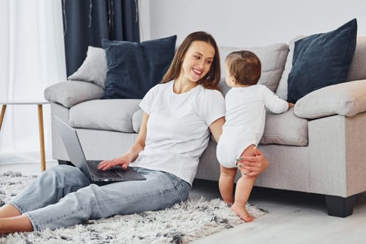 Silver colored laptop. Mother with her little daughter is indoors at home together.