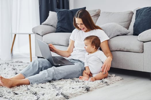 Silver colored laptop. Mother with her little daughter is indoors at home together.