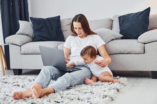 Silver colored laptop. Mother with her little daughter is indoors at home together.
