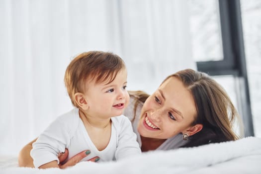 On the white bed. Mother with her little daughter is indoors at home together.