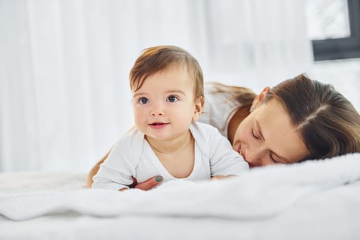 On the white bed. Mother with her little daughter is indoors at home together.