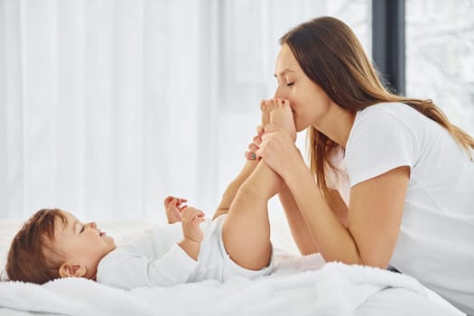 On the white bed. Mother with her little daughter is indoors at home together.