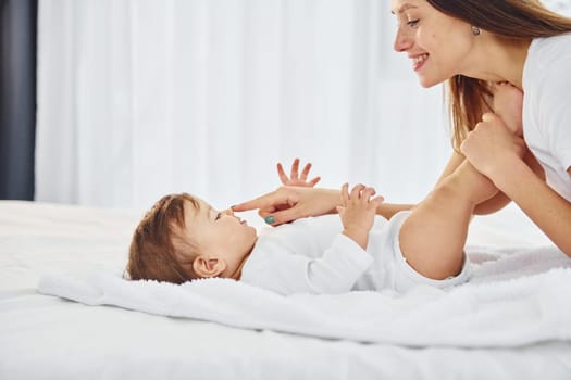 On the white bed. Mother with her little daughter is indoors at home together.