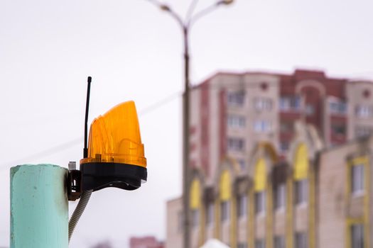 Yellow signal lamp with antenna on a metal tube. In the background, out of focus, a lamppost and residential buildings. Gloomy gray sky.