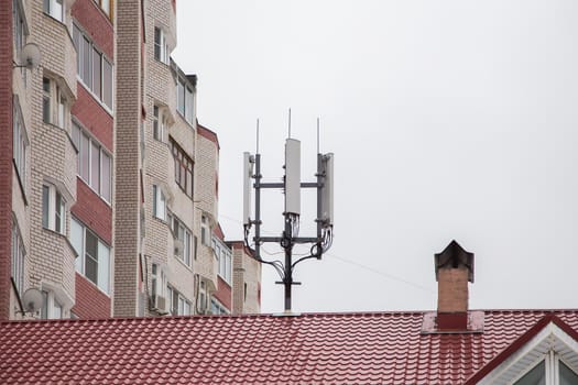 Cellular antennas on the roof next to the chimney. The device is located opposite a high-rise residential building. Against the background of the gray sky.