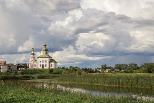 An old white-stone church on the bank of a small river. There are one-story residential buildings near the temple. Thick clouds hang from above.