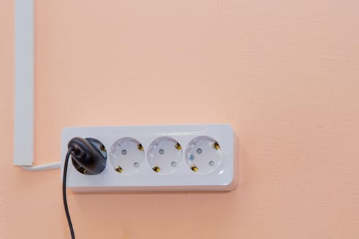 An outlet with a plugged-in plug and wires going side by side in a box. Electrical white items are fixed to the beige wall. There is copy space.