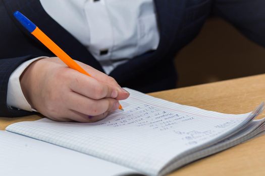 The boys hand with a fountain pen with blue ink solves math examples. The student completes the task in teradi. A schoolboy teaches lessons at a school desk. Warm soft daylight.