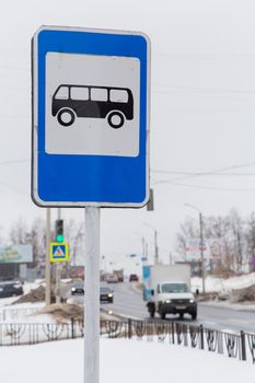 A road sign means a place to stop the bus, pick up and drop off passengers. Against the background of the road and passing cars. It's winter in the city, white snow lies on the street.