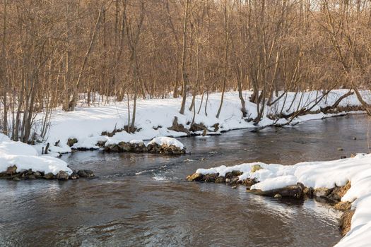 A river bed with snow on the banks and high bushes. In a narrow place of the river bed, a threshold is formed, along which water flows violently. Beautiful nature in a winter evening.