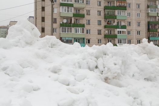 A large snowdrift in the city against the background of a multi-storey building. A block of snow with dirty lumps lies on a city street in the courtyard of a residential building.
