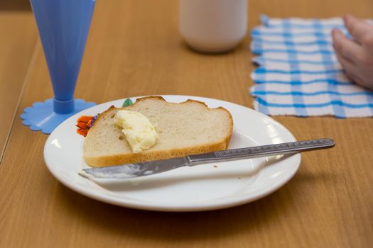 Close-up of a piece of white bread with butter and a scabbard on a white plate. A plate with a sandwich is on the table. In the background, in defocusing, a glass and a napkin.