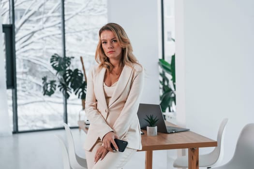 Standing near window. Business woman in formal clothes is indoors posing for the camera.