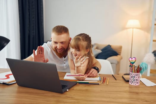 By the table with laptop on it. Father with his little daughter is at home together.