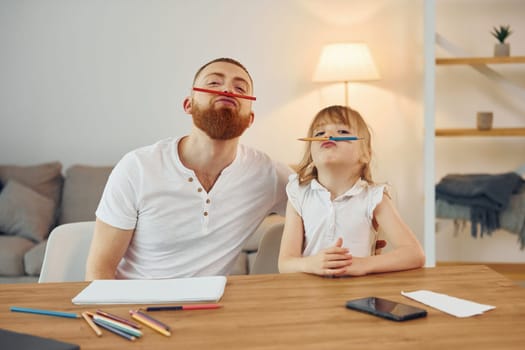 Sitting by the table and having fun. Father with his little daughter is at home together.