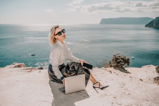 Freelance women sea working on a computer. Pretty middle aged woman with computer and phone outdoors with beautiful sea view. The concept of remote work