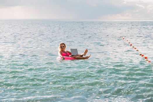 Woman works on laptop in sea. Freelancer, blond woman in sunglases floating on an inflatable big pink donut with a laptop in the sea at sunset. People summer vacation rest lifestyle concept