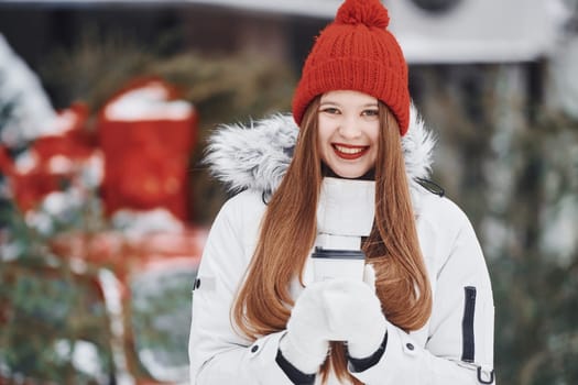 With cup of drink in hands. Happy young woman standing outdoors and celebrating christmas holidays.