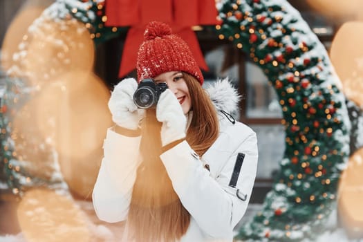 With camera in hands. Happy young woman standing outdoors and celebrating christmas holidays.