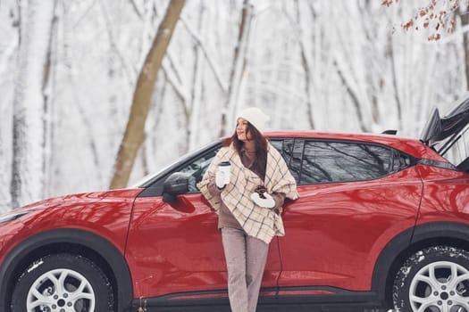 With cup of drink. Beautiful young woman is outdoors near her red automobile at winter time.