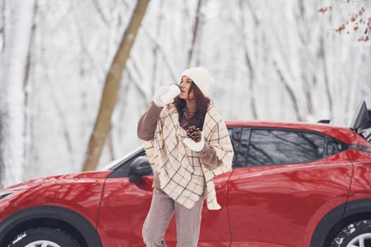 With cup of drink. Beautiful young woman is outdoors near her red automobile at winter time.