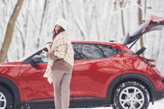 With cup of drink. Beautiful young woman is outdoors near her red automobile at winter time.