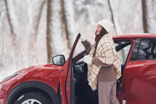 Warm drink in cup. Beautiful young woman is outdoors near her red automobile at winter time.