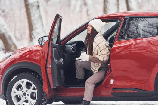 Warm drink in cup. Beautiful young woman is outdoors near her red automobile at winter time.