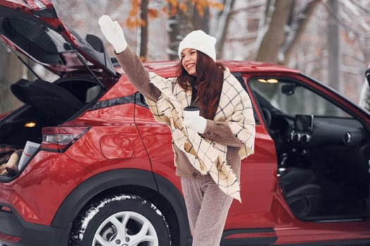 Hello, gesture. Beautiful young woman is outdoors near her red automobile at winter time.