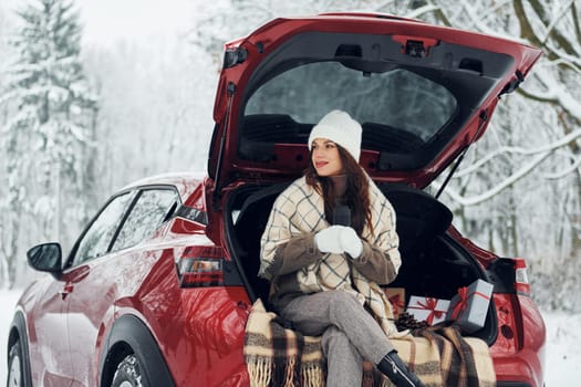 Sitting on the back of a car. Beautiful young woman is outdoors near her red automobile at winter time.