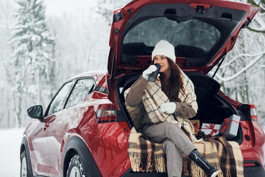 Sitting on the back of a car. Beautiful young woman is outdoors near her red automobile at winter time.
