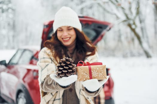 Holding gift box. Beautiful young woman is outdoors near her red automobile at winter time.