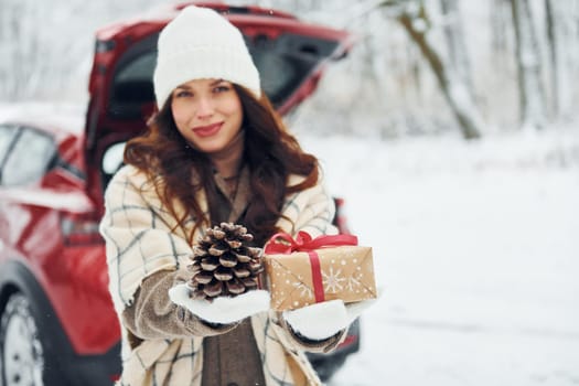 Holding gift box. Beautiful young woman is outdoors near her red automobile at winter time.