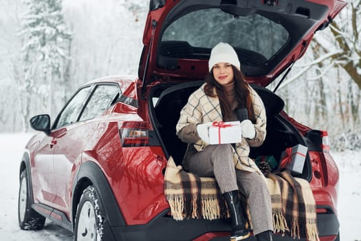 Holding gift box. Beautiful young woman is outdoors near her red automobile at winter time.
